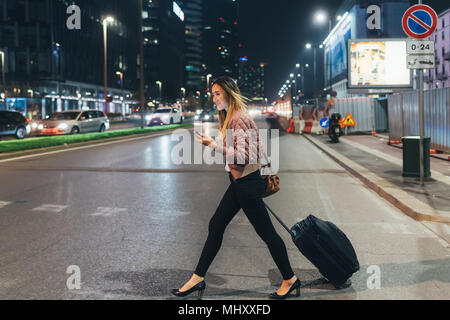 Woman walking outdoors at night, pulling wheeled suitcase, holding smartphone Stock Photo
