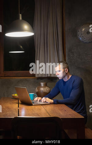 Mature man at dining table typing on laptop, night Stock Photo