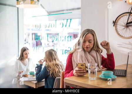 Woman sitting in cafe, looking at smartphone, laptop in front of her Stock Photo