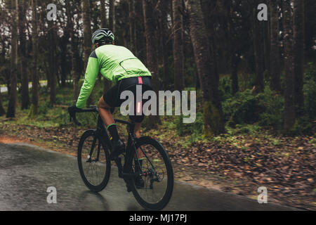 Male cyclist on a country road training for competition. Man riding bicycle on wet road through forest. Stock Photo