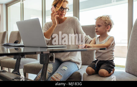 Beautiful young mother talking on mobile phone with her toddler son sitting by. Woman with son working from home. Stock Photo