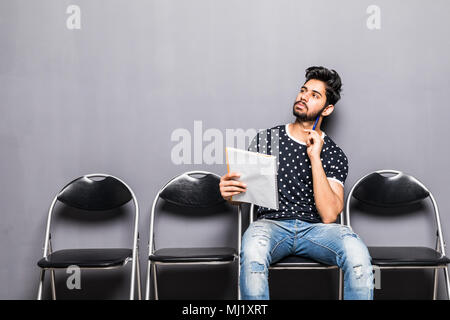 Long time of waiting. Pensive young businessman holding paper and holding hand on chin while sitting on chair against grey background Stock Photo