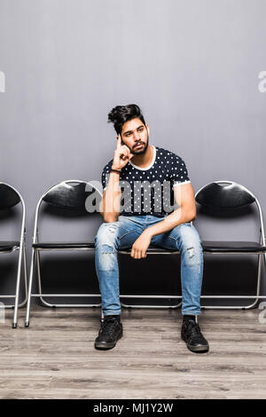 Long time of waiting. Pensive young businessman holding paper and holding hand on chin while sitting on chair against grey background Stock Photo