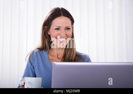 Close-up Of Happy Woman Holding Coffee Cup Looking At Laptop On White Desk Stock Photo