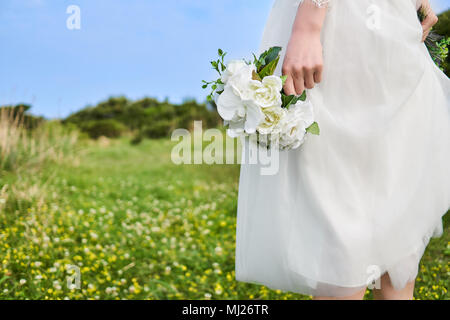 Asian woman wearing white laced bridal dress  holding white bouquet at her side at the yellow blossom field located on the cliffs of the beautiful Jej Stock Photo