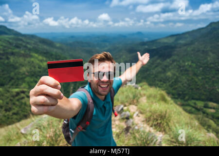 Young smiling male showing empty credit card. He is standing on top of mountain and showing the outdoor beauty. Stock Photo