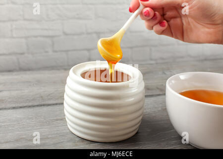 Detail on woman holding small ceramic  spoon, filled with honey, about to put it in white tea cup full of hot tea. Stock Photo