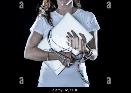 Closeup of a overworked, tired business woman holding a laptop with cables wrapped around her hands on a black background in a computer addiction conc Stock Photo
