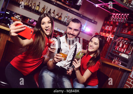 friends - two young attractive girls and a guy at a nightclub party make a photo of themselves on a mobile phone or smartphone Stock Photo