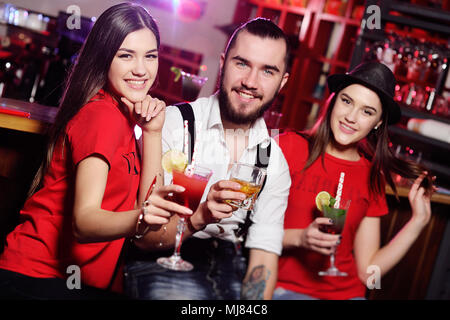 three friends - cute guy and two attractive young girls at a party holding cocktails in front of a bar smiling Stock Photo