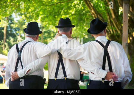 Rear view of three men wearing white shirts, black trousers with suspenders and hat standing side by side, arms around each other. Stock Photo