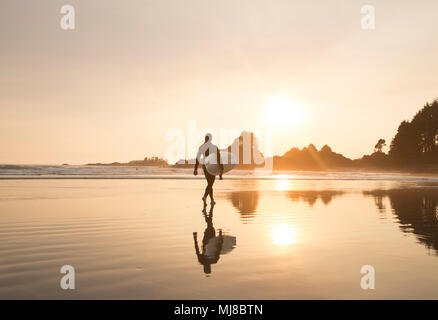Reflection of man wearing wet suit and carrying surfboard walking along sandy beach at sunset. Stock Photo