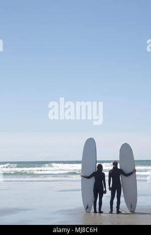Rear view of man and woman wearing wet suits standing side by side on sandy beach by the ocean, holding surfboards. Stock Photo