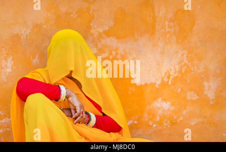 Woman wearing red and yellow sari and veil covering her head sitting on floor, leaning against orange wall. Stock Photo