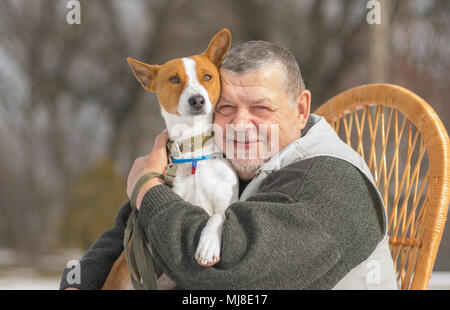 Outdoor portrait of Caucasian senior man with his cute basenji dog sitting in wicker chair at sunny day Stock Photo