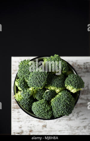 high angle view of some stems of broccoli in a white ceramic bowl, on a rustic wooden table, against a black background with some blank space on top Stock Photo