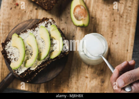 high angle view of a slice of rye bread topped with some slices of avocado, and the hand of a man taking some yoghurt from a glass jar with a spoon, o Stock Photo