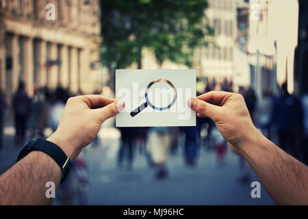 Close up of man hands holding a white paper sheet with magnifier icon inside. Searching for someone on a crowded city street background. Stock Photo