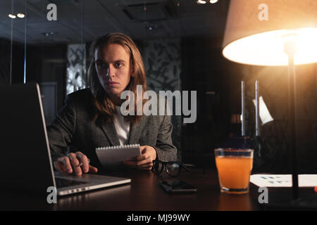 Young hipster businessman sits by the wooden table with lamp, laptop and smartphone at night office. Man with long hair work with contemporary noteboo Stock Photo