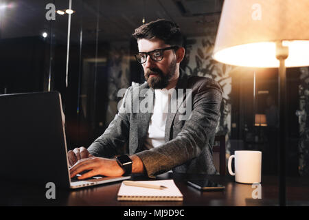 Bearded young businessman working on open space office at night. Man using modern notebook, typing text. Coworker working. Film effect Stock Photo