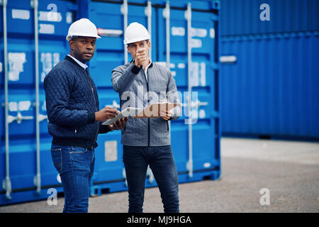 Two engineers tracking inventory using a clipboard and digital tablet while standing by freight containers on a large commercial shipping dock Stock Photo