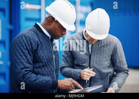 Two engineers standing together by freight containers on a large commercial shipping dock comparing inventory lists on a tablet and a clipboard Stock Photo