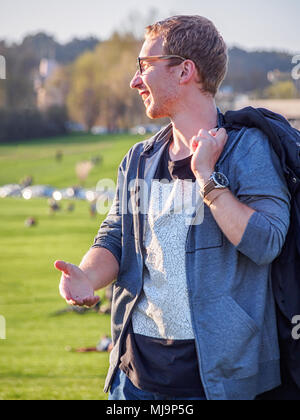 Side view of a laughing blonde white young man in glasses with wristwatch (on a background of a city park) Stock Photo