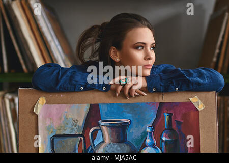 Young beautiful girl, female artist painter thinking of a new artwork idea or project holding a finished picture artwork depicting still life in blue  Stock Photo