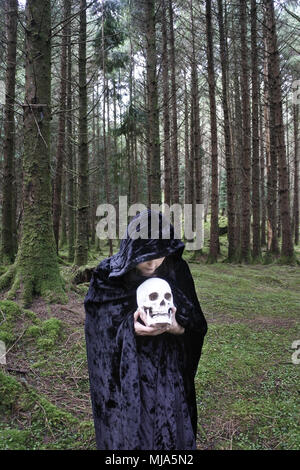 A female witch holding a human skull in dark woods - John Gollop Stock Photo