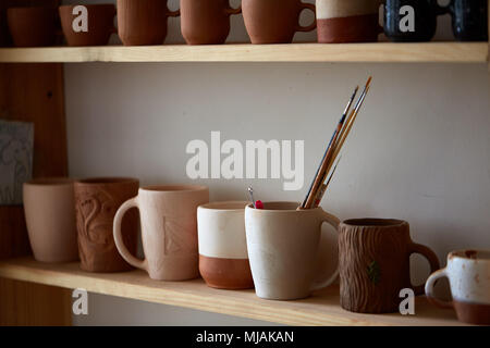 Different handmade pottery arranged in rows on a wooden shelf, close-up, selective focus. Decorative clay mugs set up on shelves over light wall. Simp Stock Photo