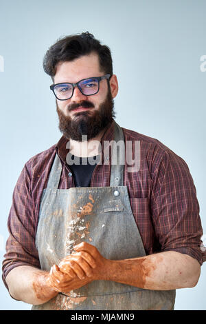 Portrait of handsome young bearded potter posing in studio, close-up. Artistic craftsman wearing plaid shirt and dirty apron holding his hands covered Stock Photo