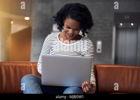 Smiling young African college student sitting alone on a campus sofa doing homework on a laptop Stock Photo