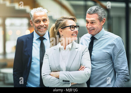 Smiling mature businesswoman talking with two male coworkers while standing together in a modern office Stock Photo