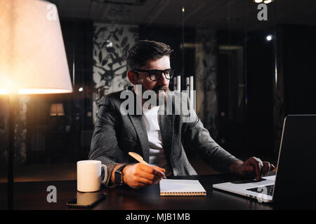 Young bearded project manager using modern laptop in loft office at night. Businessman working process. Film effect Stock Photo