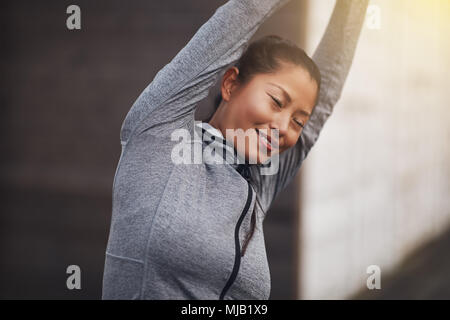 Fit young Asian woman in sportswear smiling while standing outside stretching her arms before exercising Stock Photo