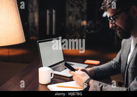 Smiling businessman with laptop and smartwatch using smartphone at night loft coworking studio. Bearded manager sits by the wooden table at modern off Stock Photo