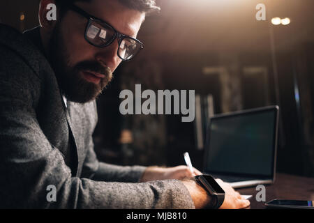 Close-up portrait of bearded man wearing eye glasses and smartwatch at night coworking loft space. Young creative manager looking away. Businessman us Stock Photo