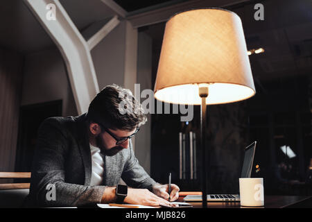 Creative businessman working with paper documents and contemporary laptop at late evening in loft office. Bearded young man sits by the wooden table w Stock Photo