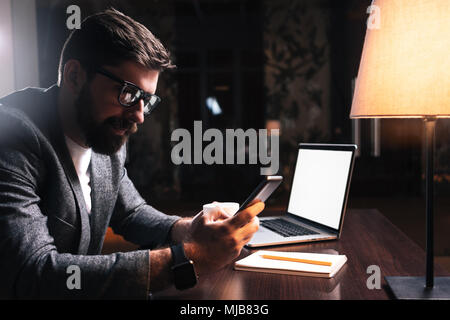 Young bearded businessman using phone while sitting by the wooden table in modern office at night. People working mobile devices. Coworking concept Stock Photo