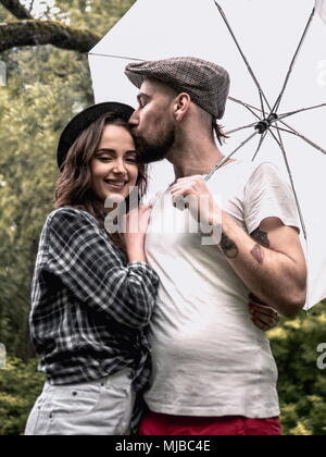 beautiful young couple man and woman in summer clothes jumping, hugging, kissing and laughing in the park with wtite umbrella under the rain Stock Photo