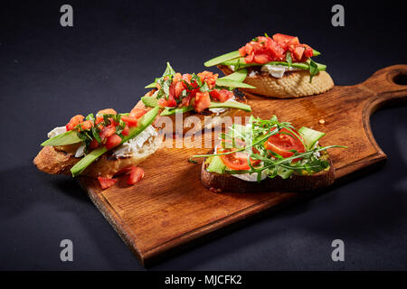 Grilled toast brisket with tomato, avocado and ruccola on wooden cutting board over dark background, top view, close-up, selective focus. Perfect vege Stock Photo