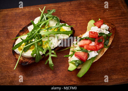 Grilled toast brisket with tomato, avocado and ruccola on wooden cutting board over dark background, top view, close-up, selective focus. Perfect vege Stock Photo