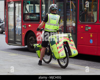 NHS London Ambulance Paramedic on a bicycle in London with hand on radio, UK. 2018. Rear view in front of red London bus. Stock Photo