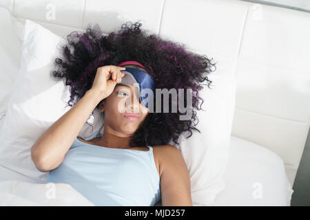 Lazy black girl with blindfold in bed late in the morning. Tired African American woman with sleeping mask sleeping during daytime. Stock Photo