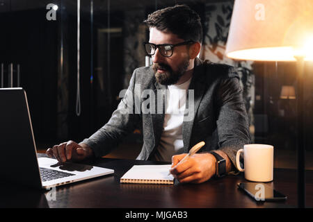 Bearded coworker sits by the wooden table with lamp and using contemporary notebook. Businessman hold pen and analysing trends. Young man working in l Stock Photo