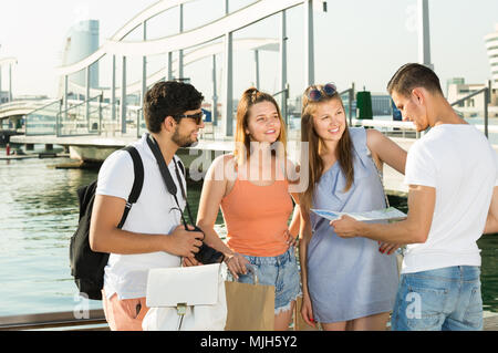 Four smiling traveling young people searching for direction using paper map on waterfront in town on summer day Stock Photo
