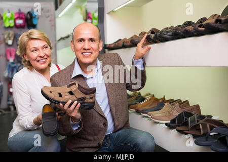 portrait of cheerful smiling mature man and woman holding picked pair of men's sandals in boutique. Focus on man Stock Photo