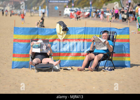 Boscombe, Bournemouth, Dorset, UK, 6th May 2018: Couple sitting on chairs with their feet up reading newspapers and relaxing on the beach in hot sunshine protected by a wind break. Hottest Mayday bank holiday weekend on record. Alamy News