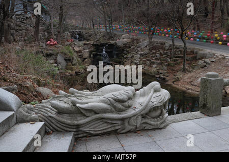 A very handsome stone dragon protects a bridge over a stream in the woods at the Donghwasa Temple in Korea; colorful lanterns line the road to temple. Stock Photo