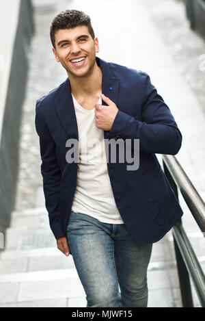 Handsome young man smiling with blue eyes in the street. Model of fashion walking in urban background wearing white t-shirt, jeans and blue jacket Stock Photo